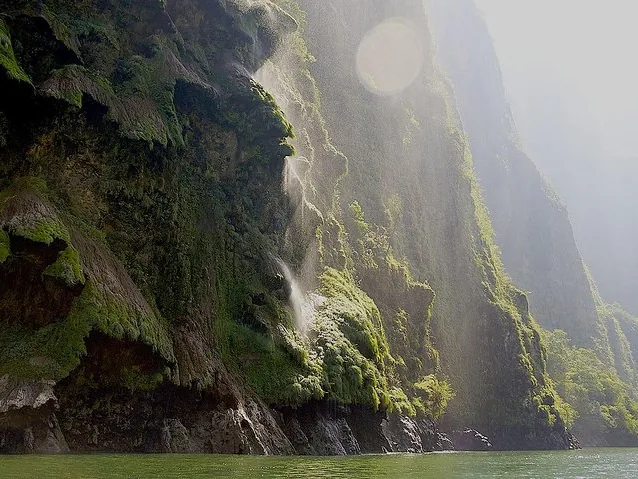 A misty waterfall cascading down a steep, moss-covered cliff into the river below